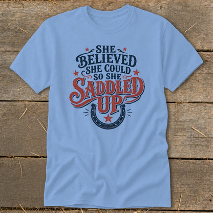 Light blue t-shirt with "She believed she could so she saddled up" text and horseshoe.