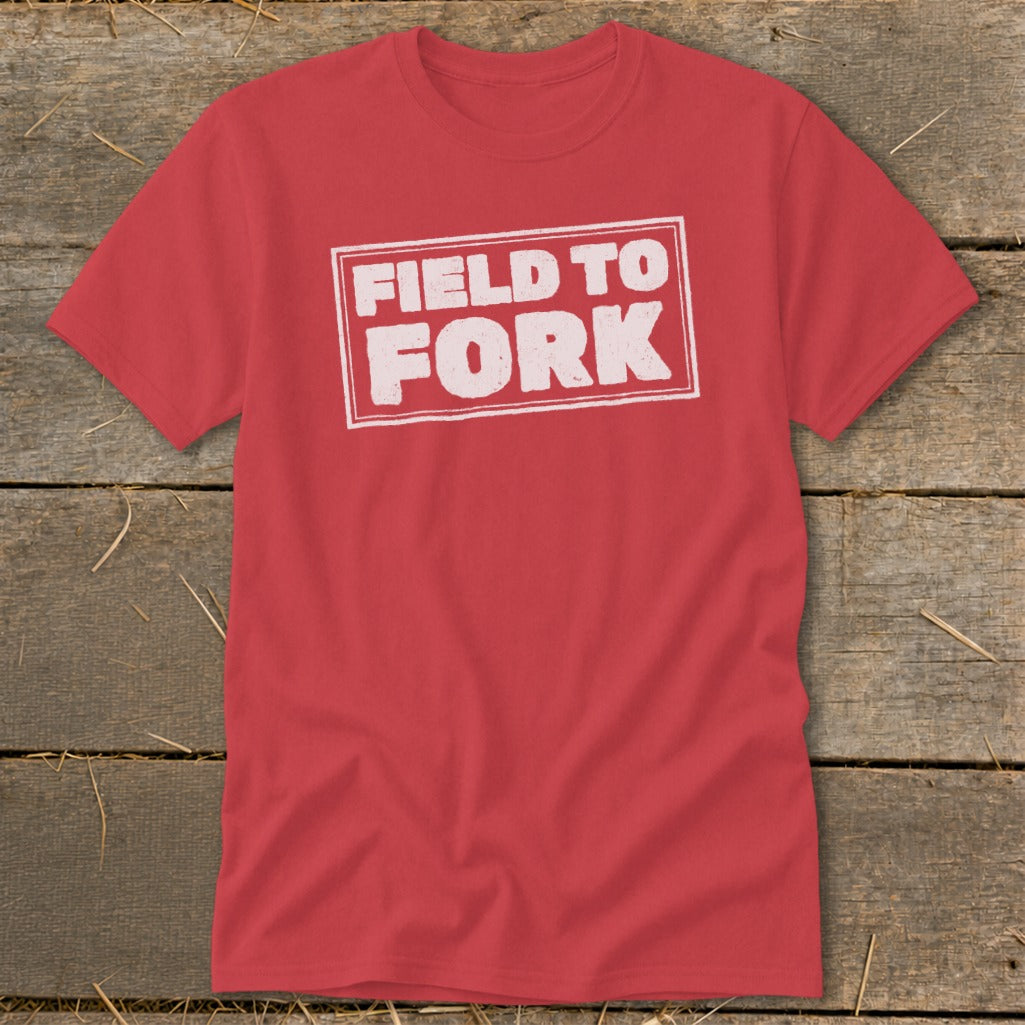 A red t-shirt with a white distressed rectangle graphic that reads FIELD TO FORK, laid flat on a rustic wooden background.