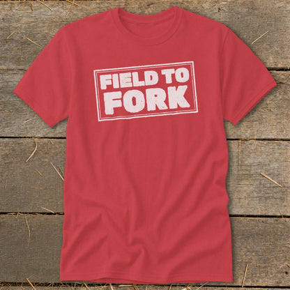 A red t-shirt with a white distressed rectangle graphic that reads FIELD TO FORK, laid flat on a rustic wooden background.