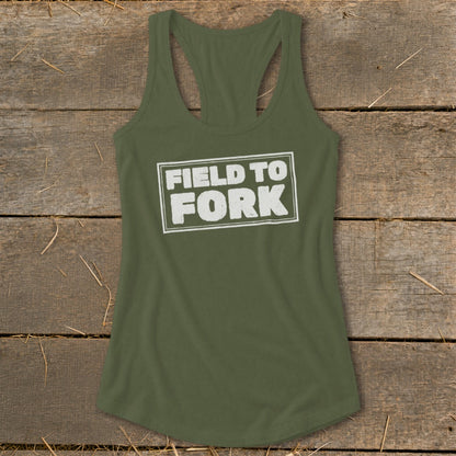 An olive green women's racerback tank top featuring the white text "FIELD TO FORK" centered on a wooden background.