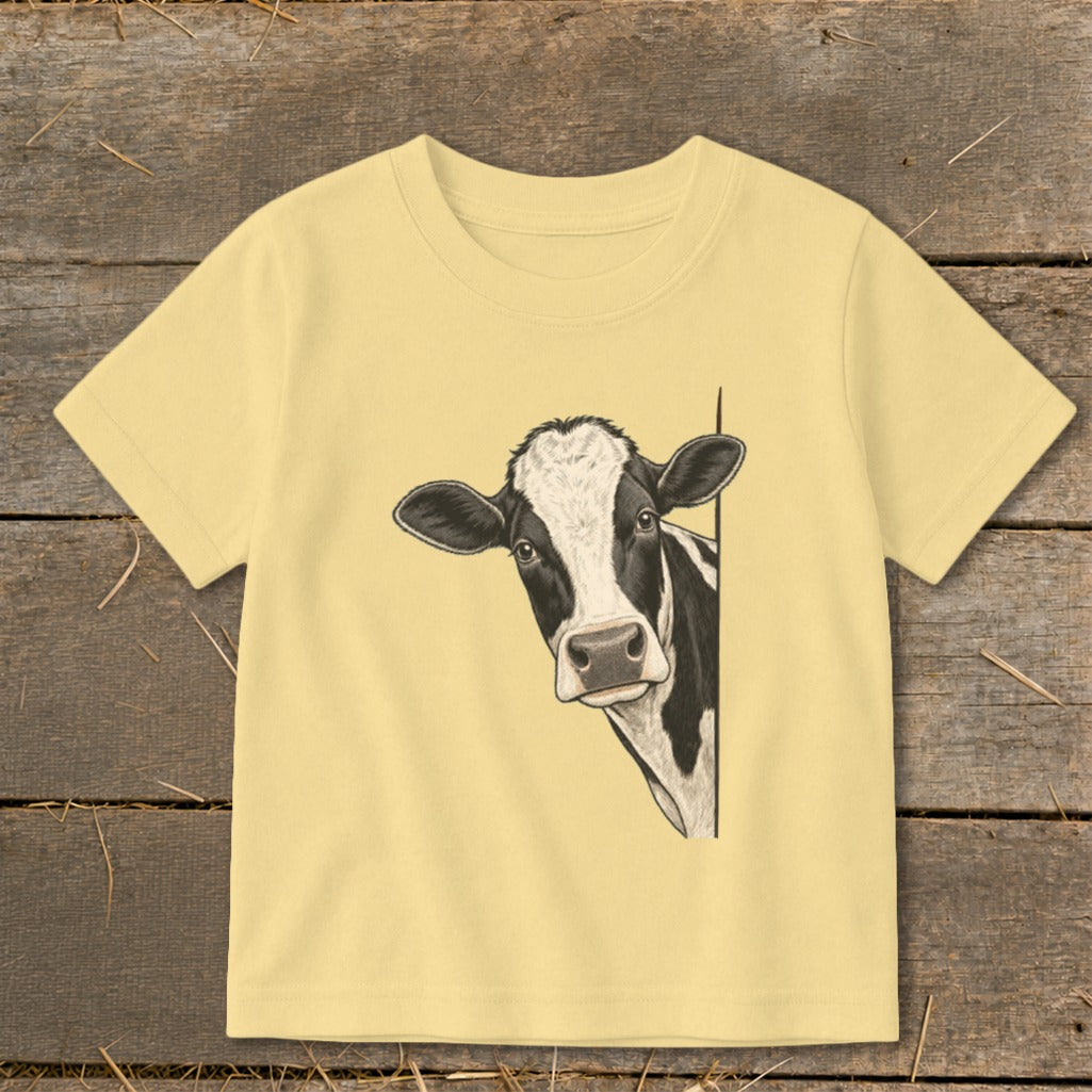 Black and white cow peeking from behind a corner on a light yellow t-shirt.