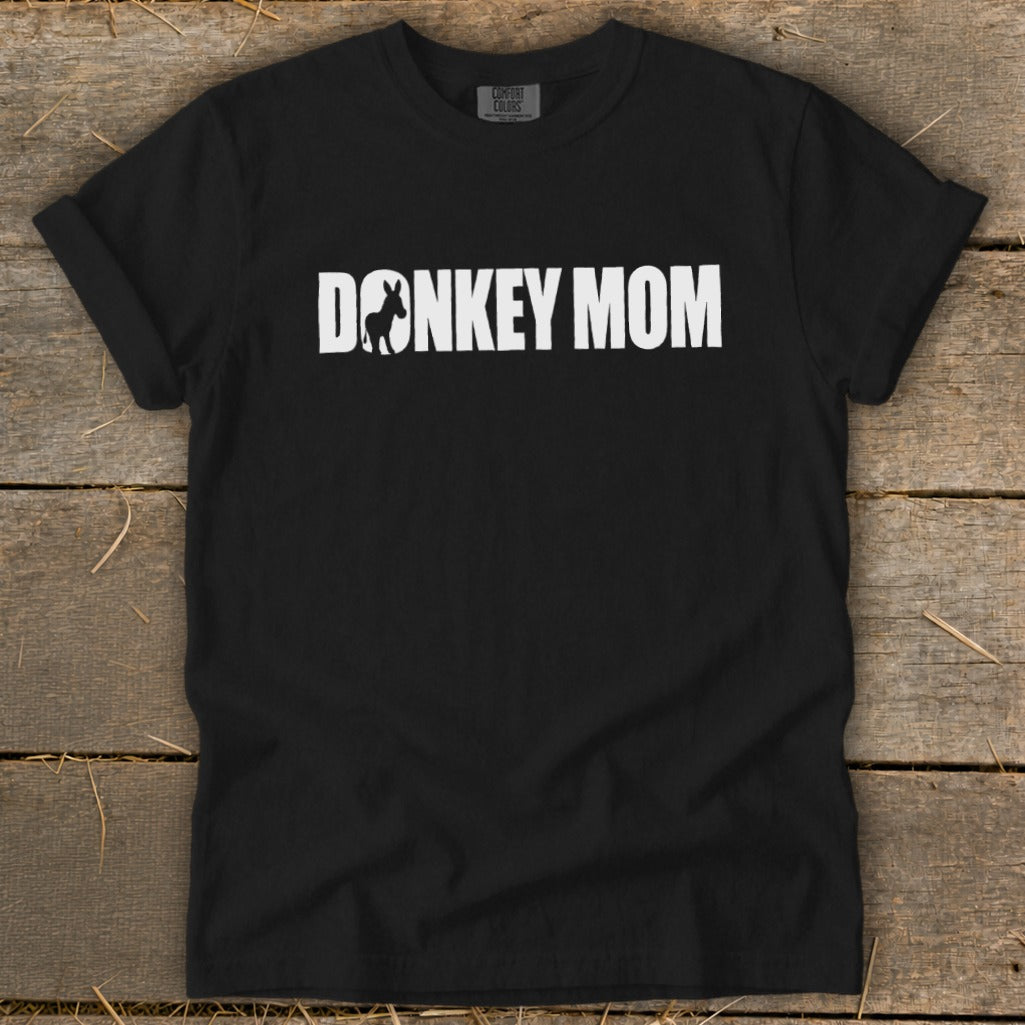 A black short-sleeved t-shirt with white text reading DONKEY MOM, featuring a donkey silhouette inside the letter O, laid on a wooden surface.