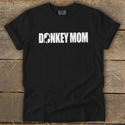 A black short-sleeved t-shirt with white text reading DONKEY MOM, featuring a donkey silhouette inside the letter O, laid on a wooden surface.