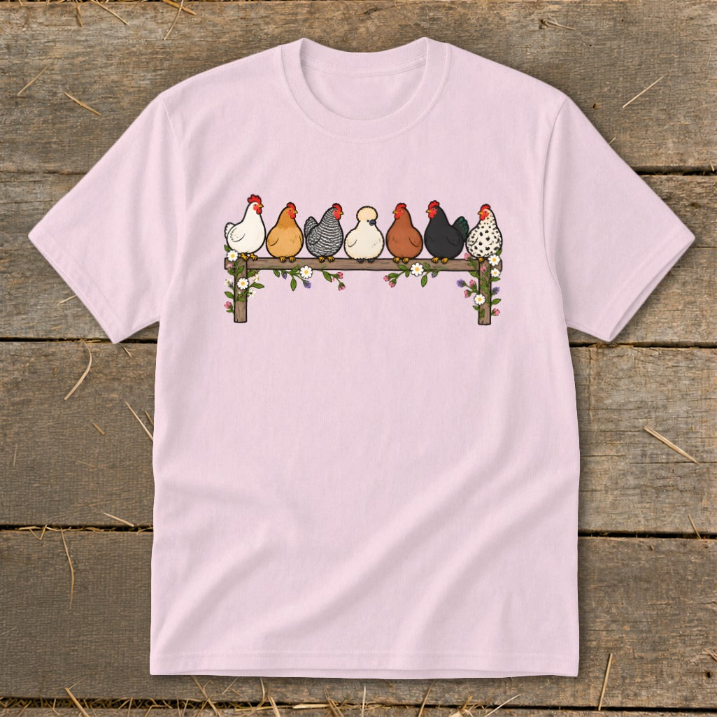 Light pink short-sleeved t-shirt featuring a cartoon graphic of seven colorful chickens roosting on a wooden fence adorned with flowers.