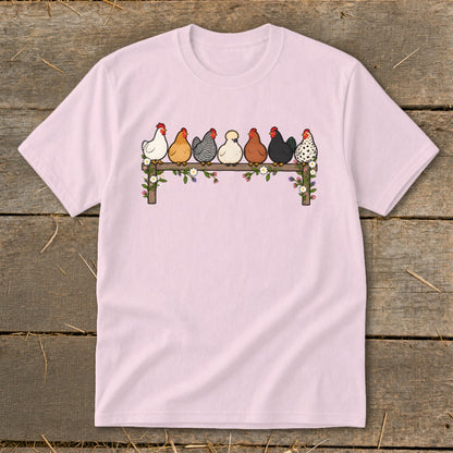 Light pink short-sleeved t-shirt featuring a cartoon graphic of seven colorful chickens roosting on a wooden fence adorned with flowers.