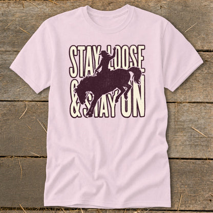 Pink t-shirt with a distressed graphic of a rodeo rider on a bucking bronco and text "STAY LOOSE & STAY ON".