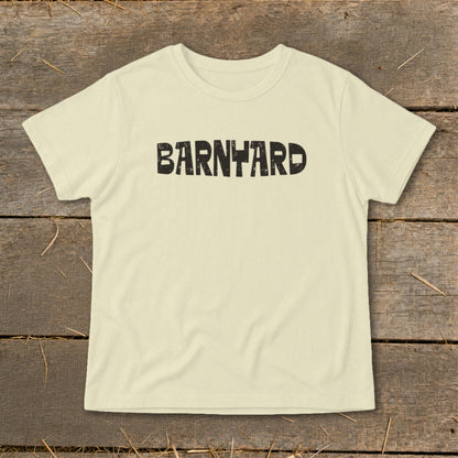 Light cream short-sleeved t-shirt featuring the word BARNYARD in distressed black block letters across the chest, displayed on a rustic wooden background.