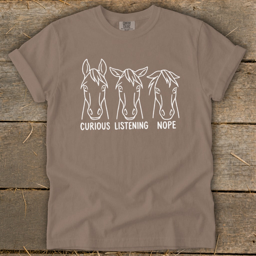 A brown crewneck t-shirt featuring a white line graphic of three horse heads with text reading CURIOUS, LISTENING, NOPE.