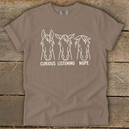 A brown crewneck t-shirt featuring a white line graphic of three horse heads with text reading CURIOUS, LISTENING, NOPE.