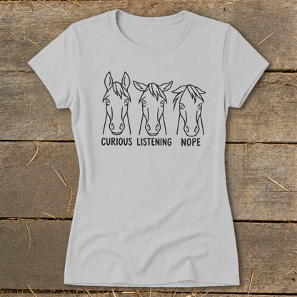 A light gray women's t-shirt features three line-drawn horse heads with the text "CURIOUS LISTENING NOPE" underneath.