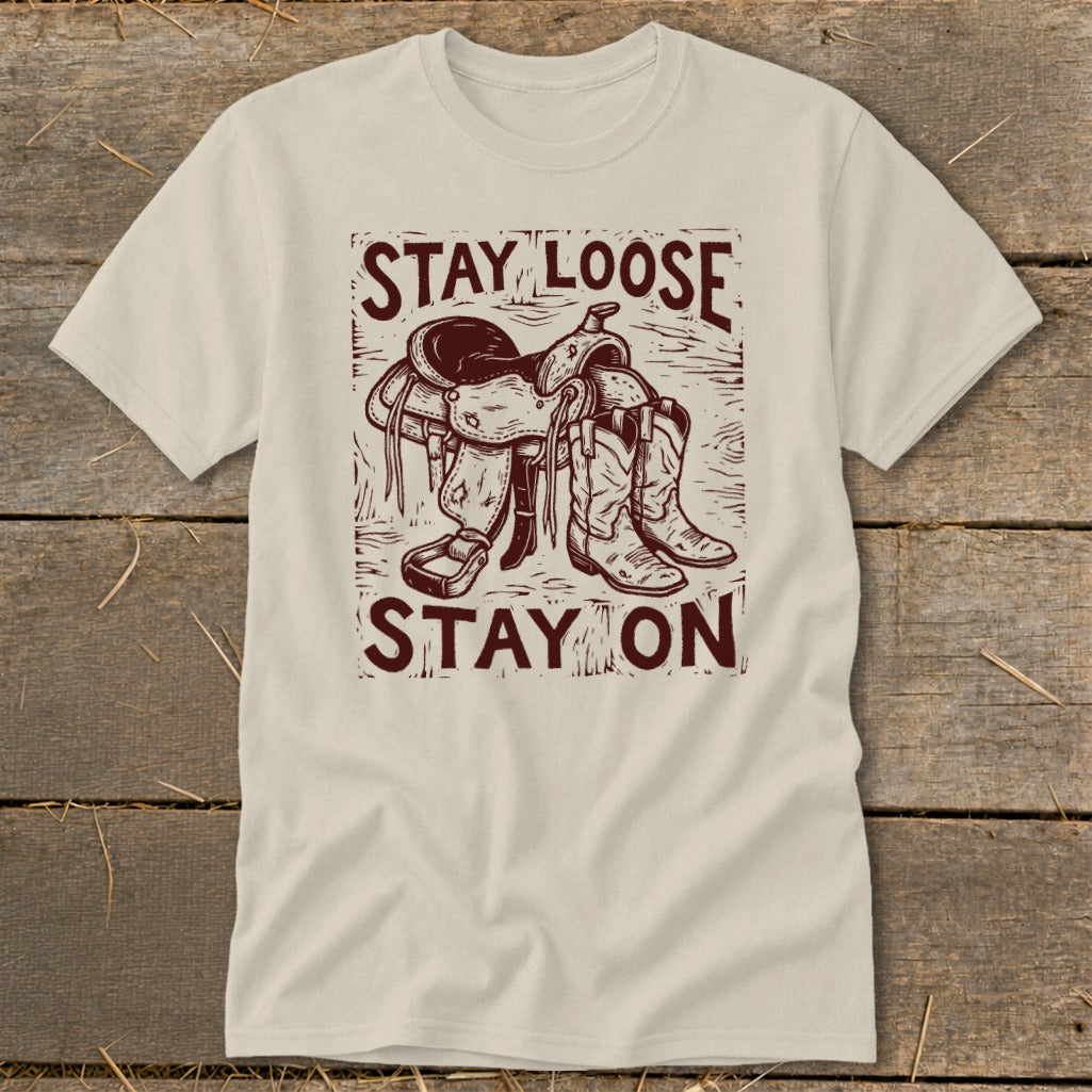 Cream t-shirt with maroon graphic of saddle and cowboy boots, saying "STAY LOOSE STAY ON".