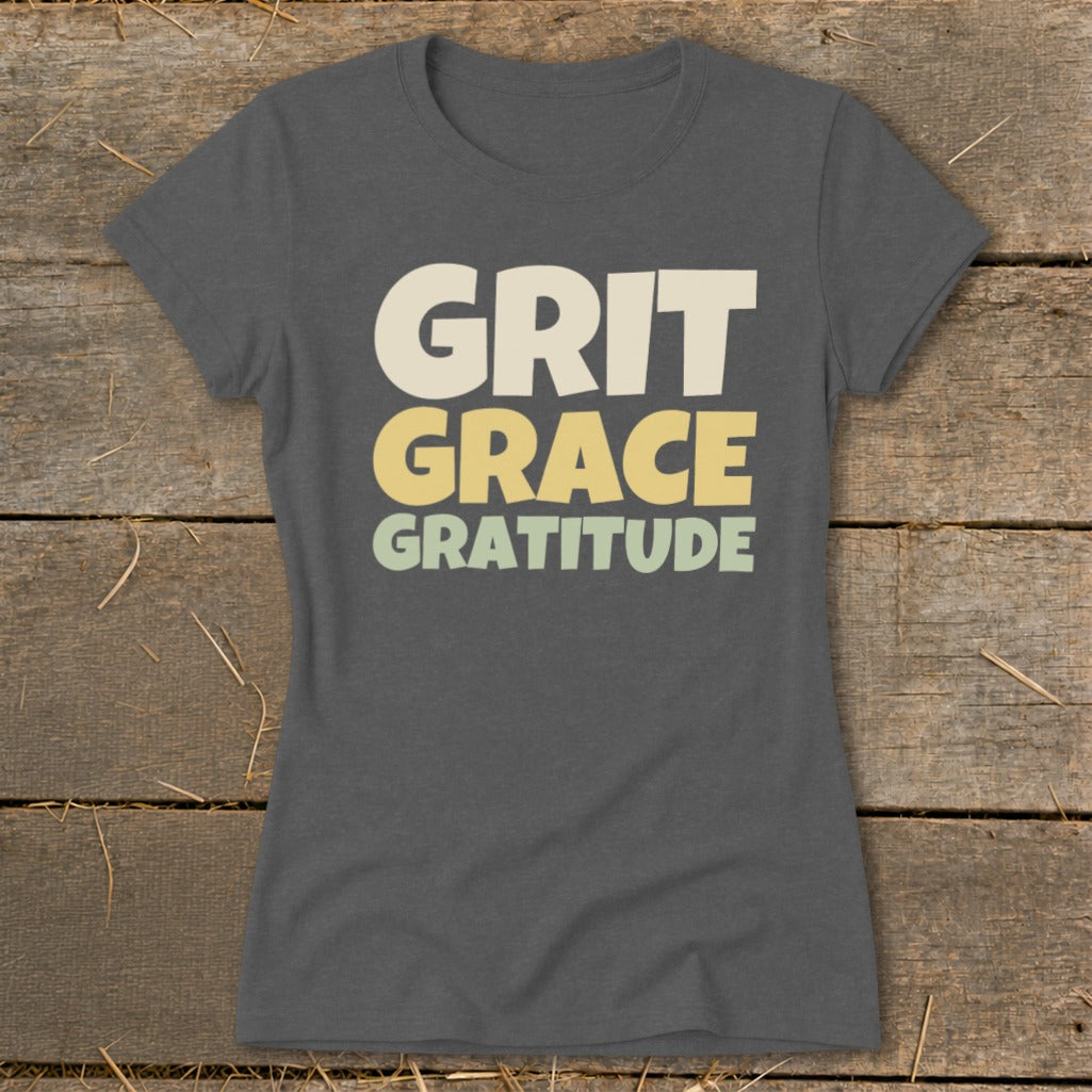 A dark gray women's t-shirt printed with the stacked text Grit Grace Gratitude in vintage-style colors on a rustic wooden background.