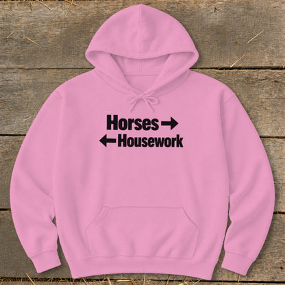 A pink hoodie lying flat on a wooden surface, featuring black text that reads Horses with a right arrow and Housework with a left arrow.