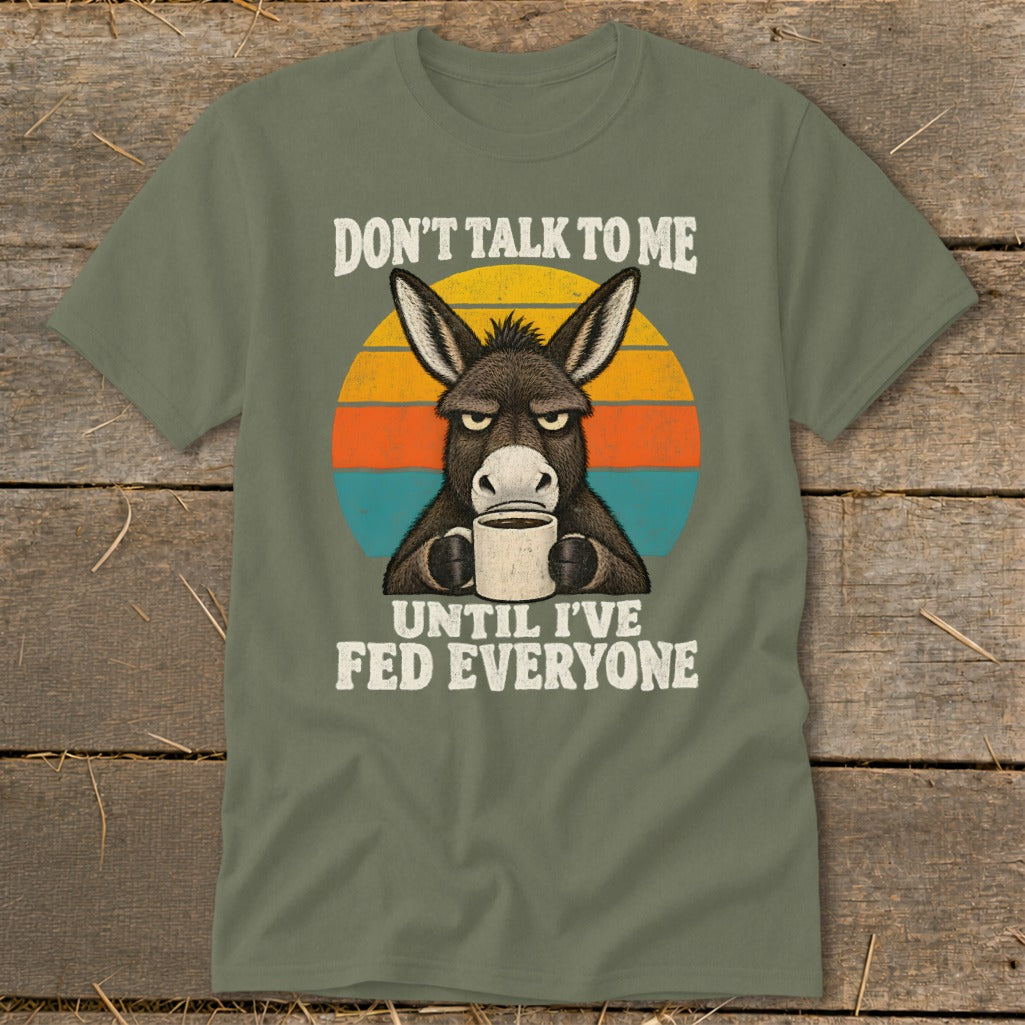 An olive green t-shirt featuring a grumpy donkey holding a coffee mug and text reading "DON'T TALK TO ME UNTIL I'VE FED EVERYONE."