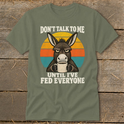 An olive green t-shirt featuring a grumpy donkey holding a coffee mug and text reading "DON'T TALK TO ME UNTIL I'VE FED EVERYONE."