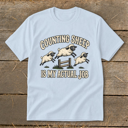 Light blue t-shirt with cartoon sheep jumping over a fence and text "Counting Sheep Is My Actual Job".
