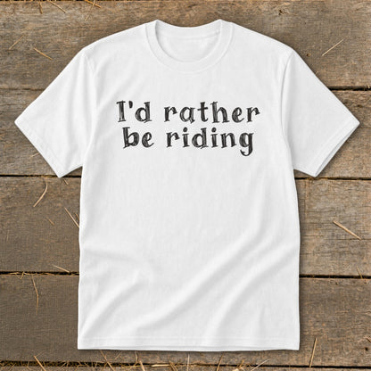 White t-shirt with black text "I'd rather be riding" on rustic wood.
