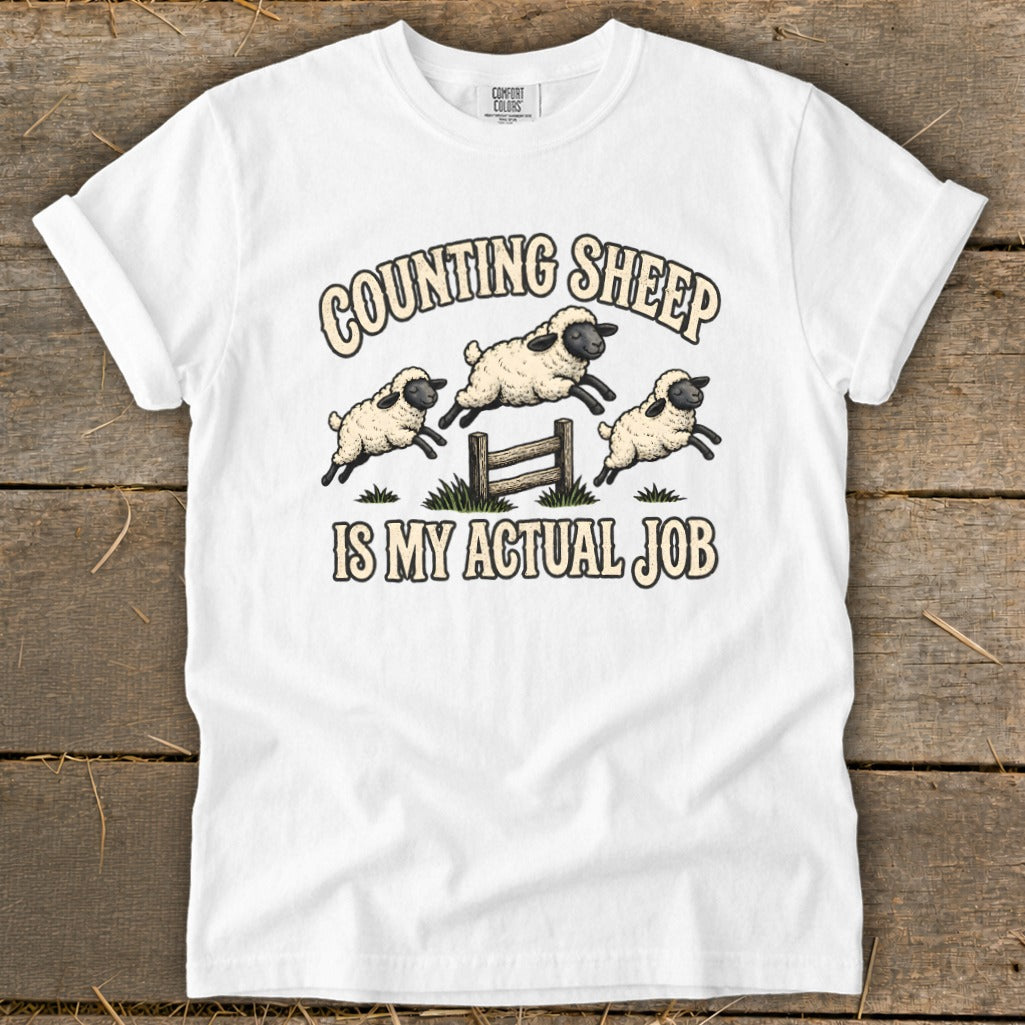 White t-shirt with cartoon sheep jumping over a fence with text "Counting Sheep Is My Actual Job".