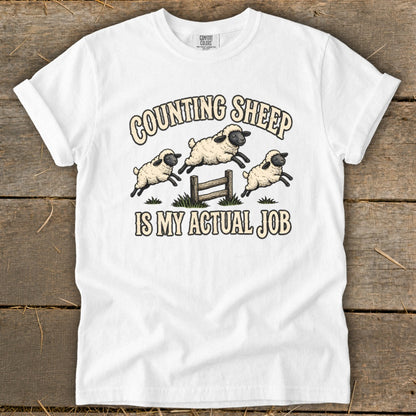 White t-shirt with cartoon sheep jumping over a fence with text "Counting Sheep Is My Actual Job".