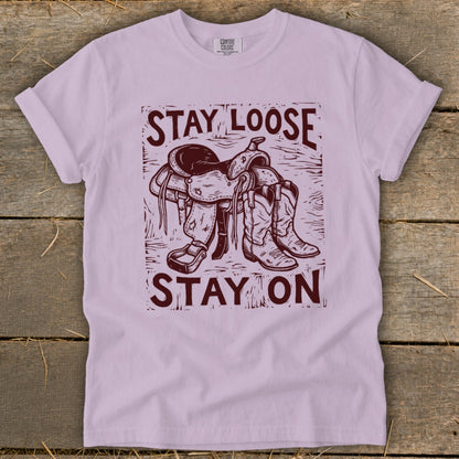 T-shirt with maroon graphic of a saddle and cowboy boots with text "STAY LOOSE STAY ON".