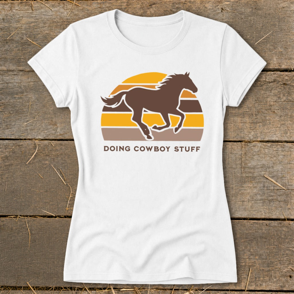 White t-shirt with a running brown horse silhouette over a striped sunset and the text "DOING COWBOY STUFF".