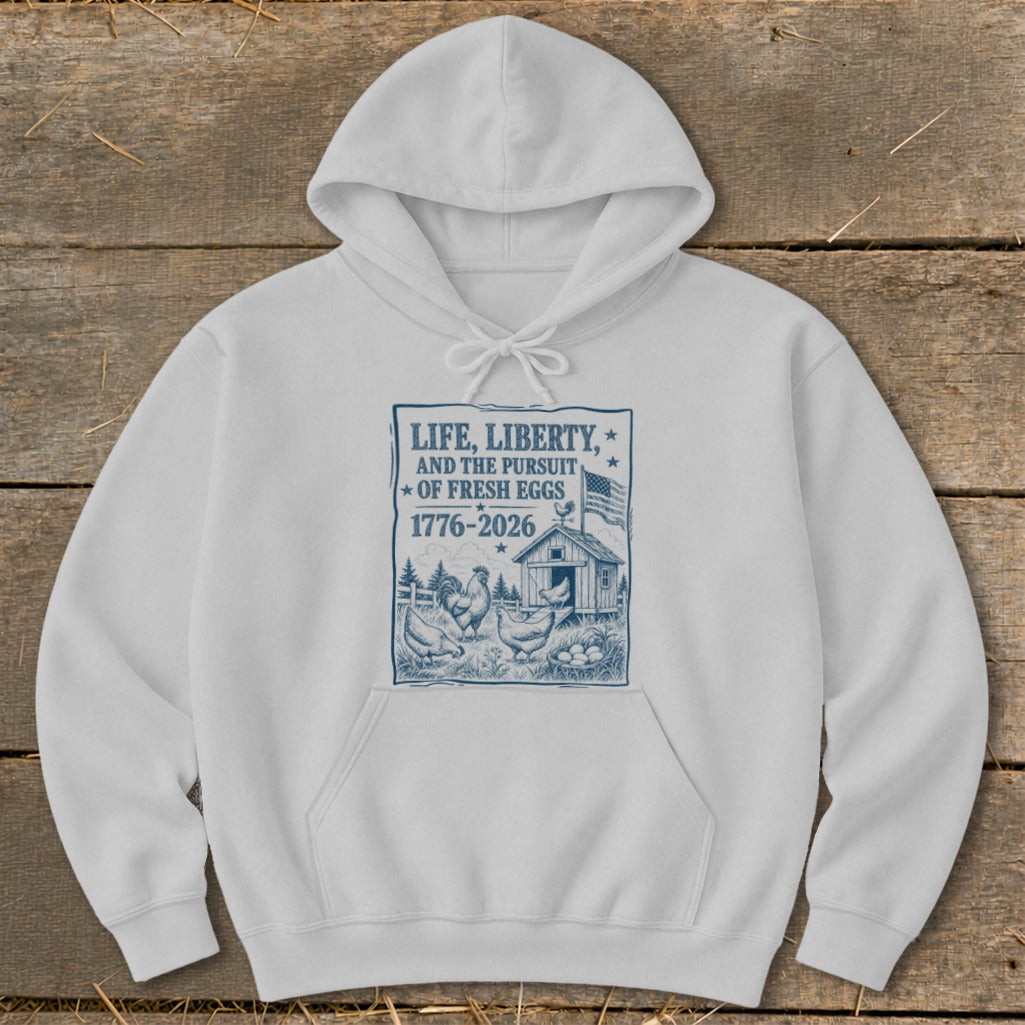 White hoodie with blue print of chickens, a coop, eggs, and text "LIFE, LIBERTY, AND THE PURSUIT OF FRESH EGGS 1776-2026".