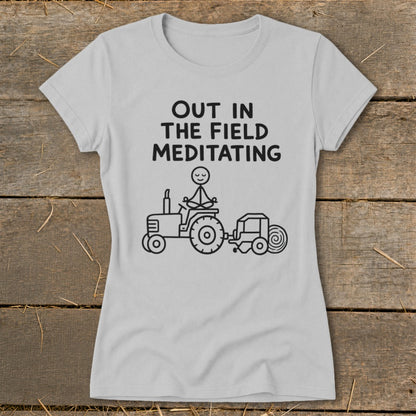 Light grey t-shirt with black text "OUT IN THE FIELD MEDITATING" and a stick figure meditating on a tractor.