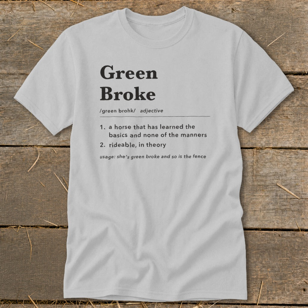 A light gray t-shirt with black text defining the term green broke laid flat on wood.