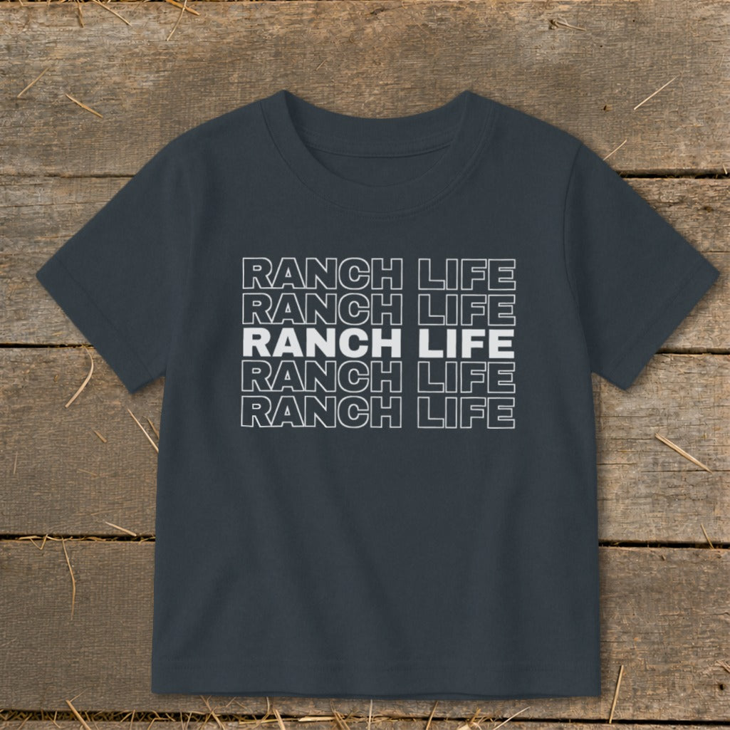 A dark gray children's t-shirt printed with the repeated white text Ranch Life on a rustic wooden background.