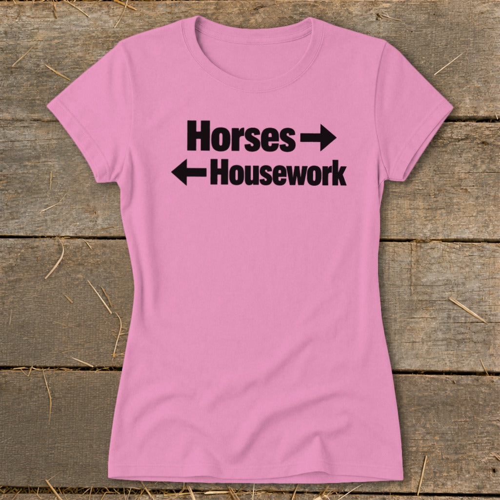 A pink women's t-shirt rests on a wooden surface, featuring black text that reads "Horses" with a right-pointing arrow above "Housework" with a left-pointing arrow.