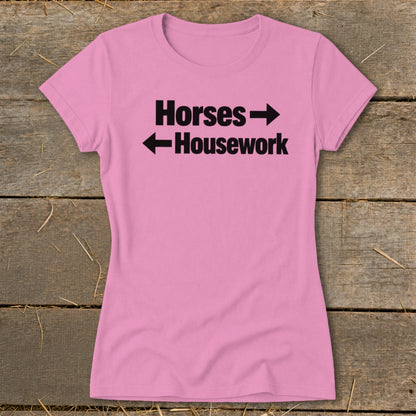A pink women's t-shirt rests on a wooden surface, featuring black text that reads "Horses" with a right-pointing arrow above "Housework" with a left-pointing arrow.