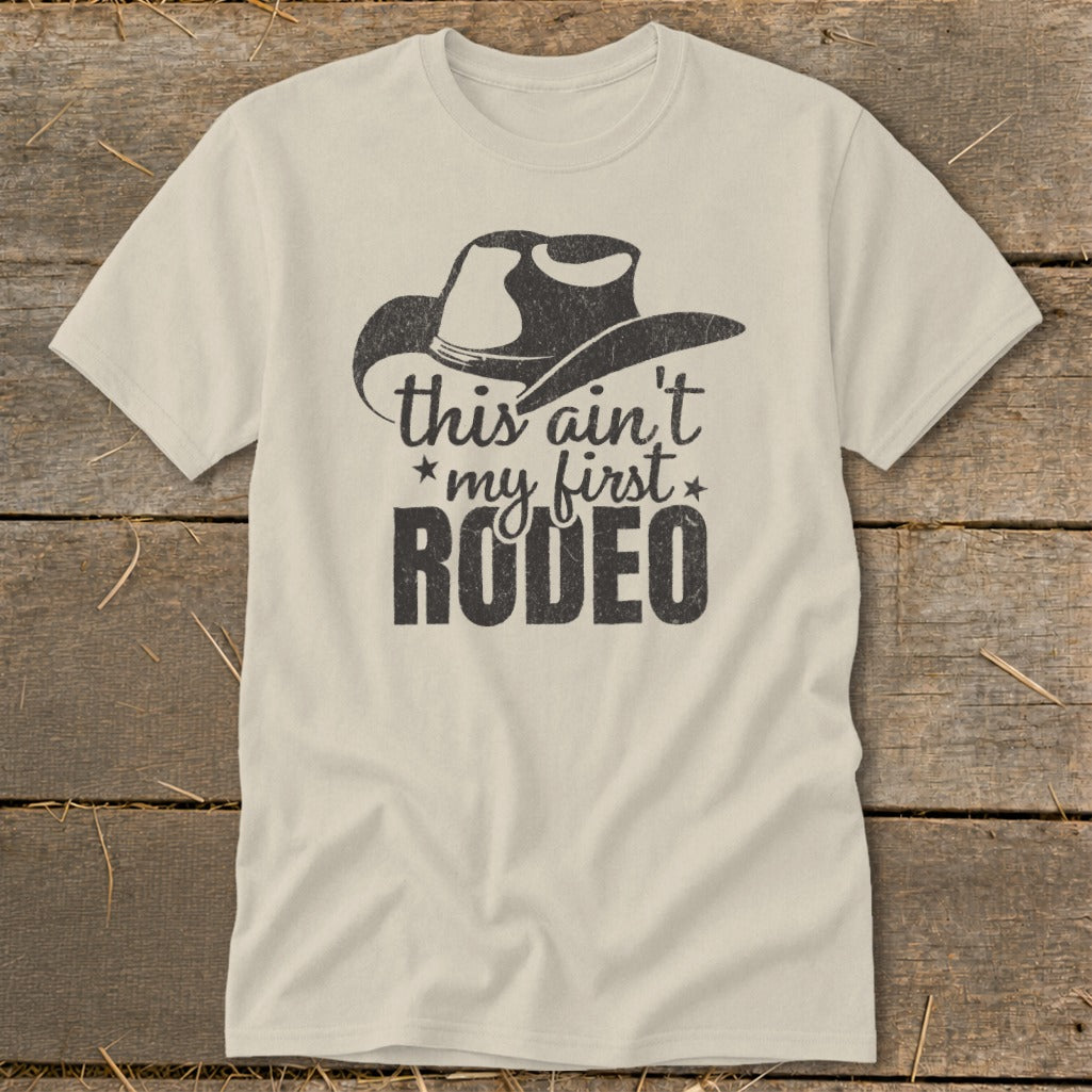 Light tan t-shirt with distressed black graphic: cowboy hat and text "this ain't my first RODEO".