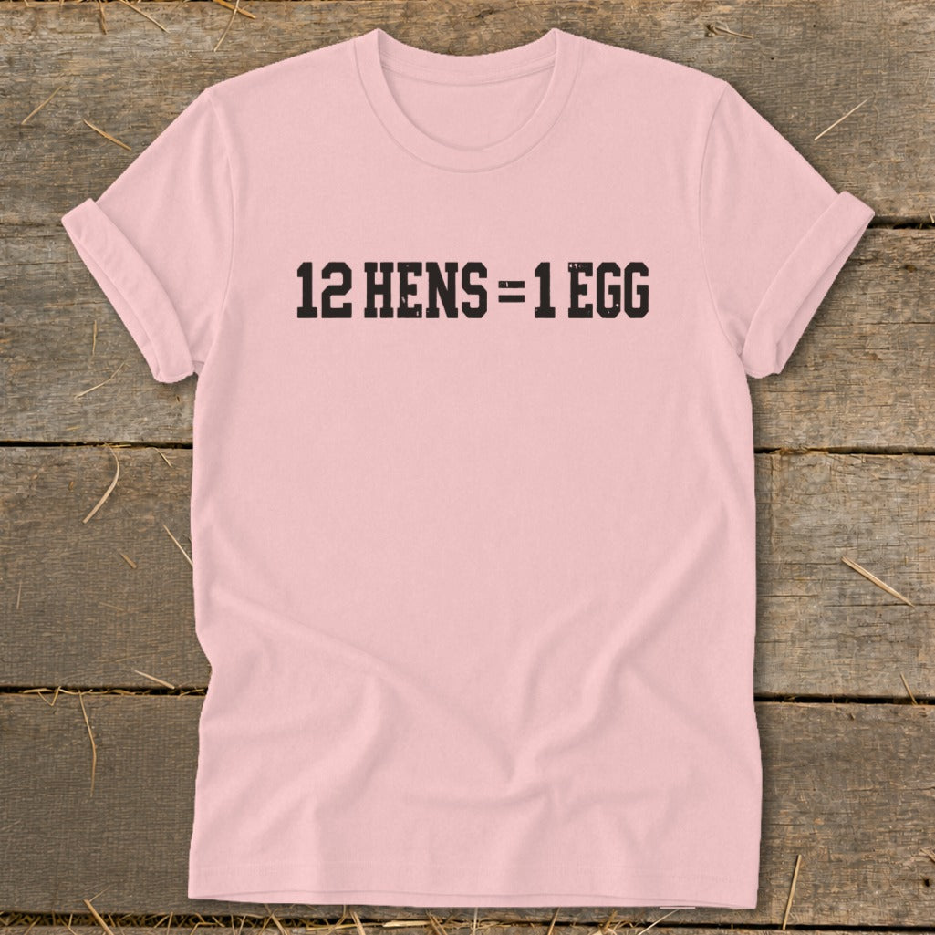 A light pink t-shirt with black text reading "12 HENS = 1 EGG" laid flat on wood.