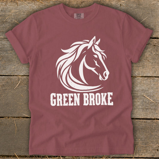 A dusty rose t-shirt lies flat on a wooden surface, featuring a white graphic of a horse head and the text "GREEN BROKE".