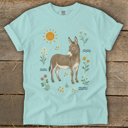 Light blue t-shirt with a watercolor illustration of a donkey surrounded by flowers, sun, and water.