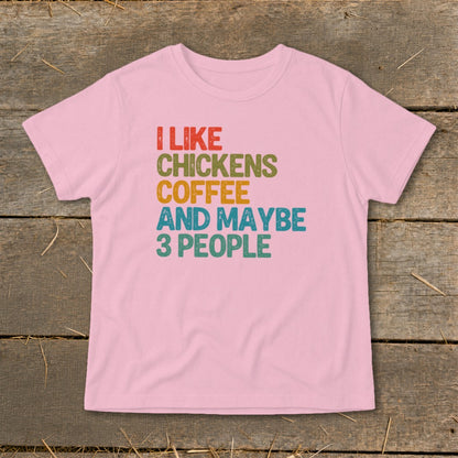 A pink short-sleeved t-shirt with distressed multicolored text reading I LIKE CHICKENS COFFEE AND MAYBE 3 PEOPLE on a wooden background.