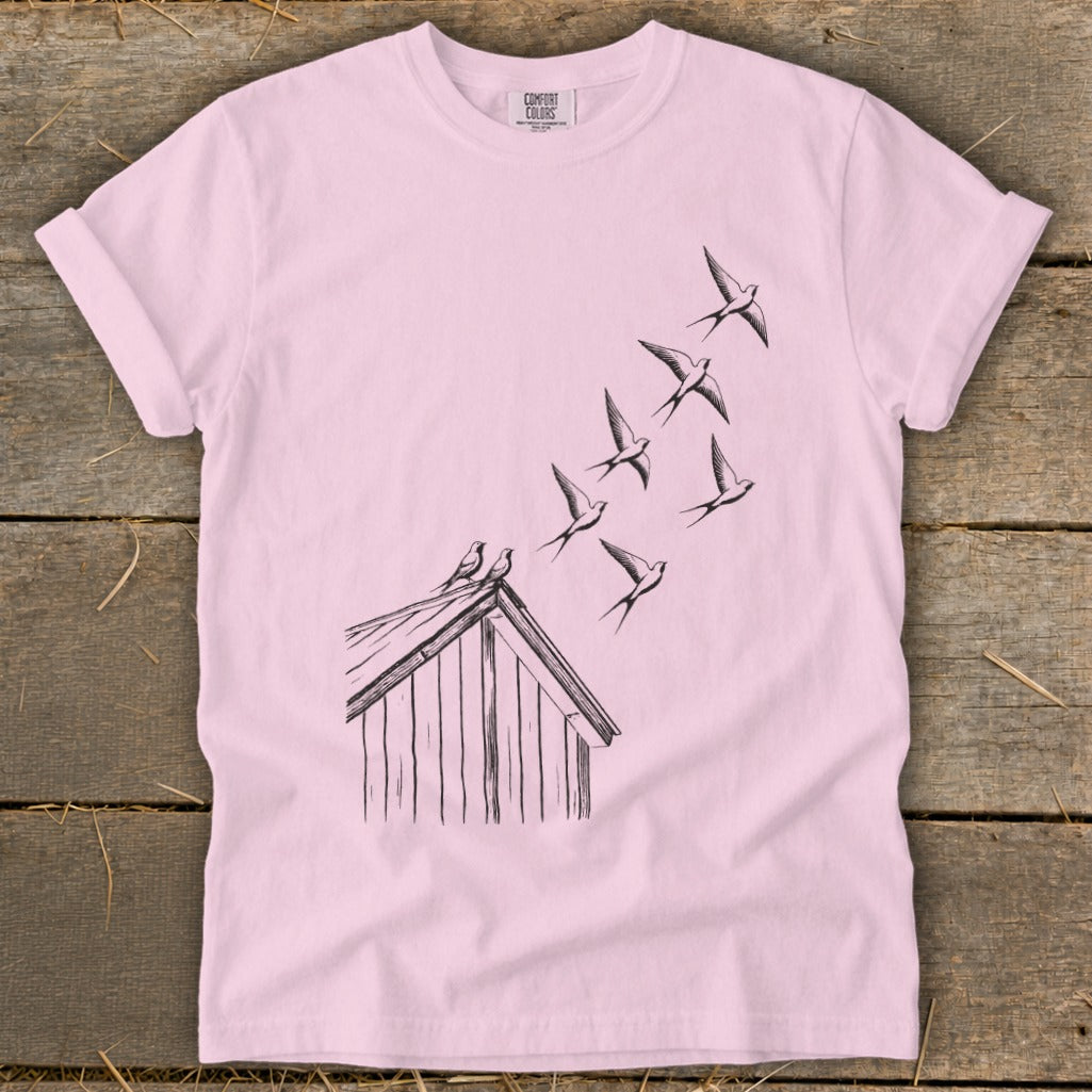 Light pink crewneck t-shirt featuring a black line drawing of birds flying from a small wooden shed.