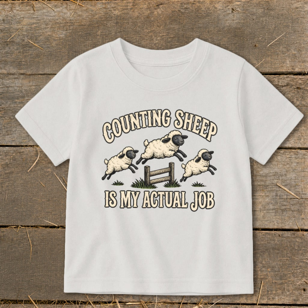 White t-shirt with cartoon sheep jumping over a fence, text reads "Counting Sheep Is My Actual Job".