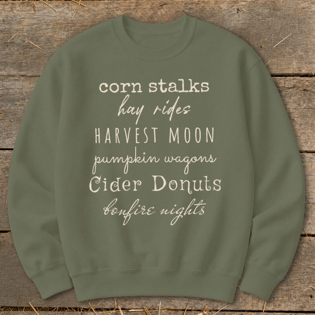 Olive green sweatshirt with white text listing fall activities: corn stalks, hay rides, harvest moon, pumpkin wagons, cider donuts, bonfire nights.
