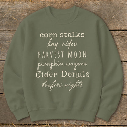 Olive green sweatshirt with white text listing fall activities: corn stalks, hay rides, harvest moon, pumpkin wagons, cider donuts, bonfire nights.