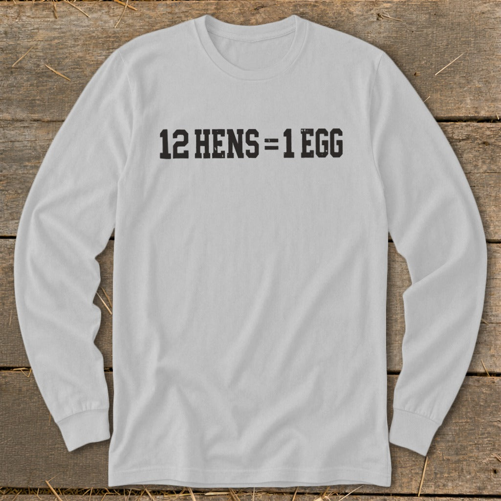 A light gray long-sleeve t-shirt with the black text 12 HENS = 1 EGG printed on the chest, laid flat on a rustic wooden background.