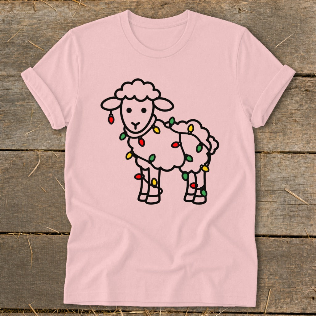 Light pink t-shirt featuring a cartoon sheep wrapped in colorful Christmas lights, laid flat on wooden planks.