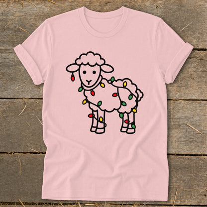 Light pink t-shirt featuring a cartoon sheep wrapped in colorful Christmas lights, laid flat on wooden planks.