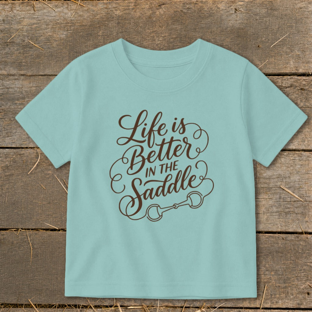 Light blue t-shirt with brown text "Life is Better IN THE Saddle" and horse bit graphic.
