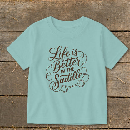 Light blue t-shirt with brown text "Life is Better IN THE Saddle" and horse bit graphic.