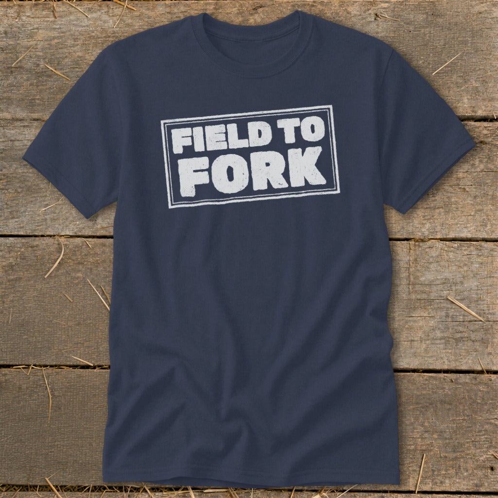 Dark blue crewneck t-shirt with white distressed text reading FIELD TO FORK centered on a rustic wooden background.