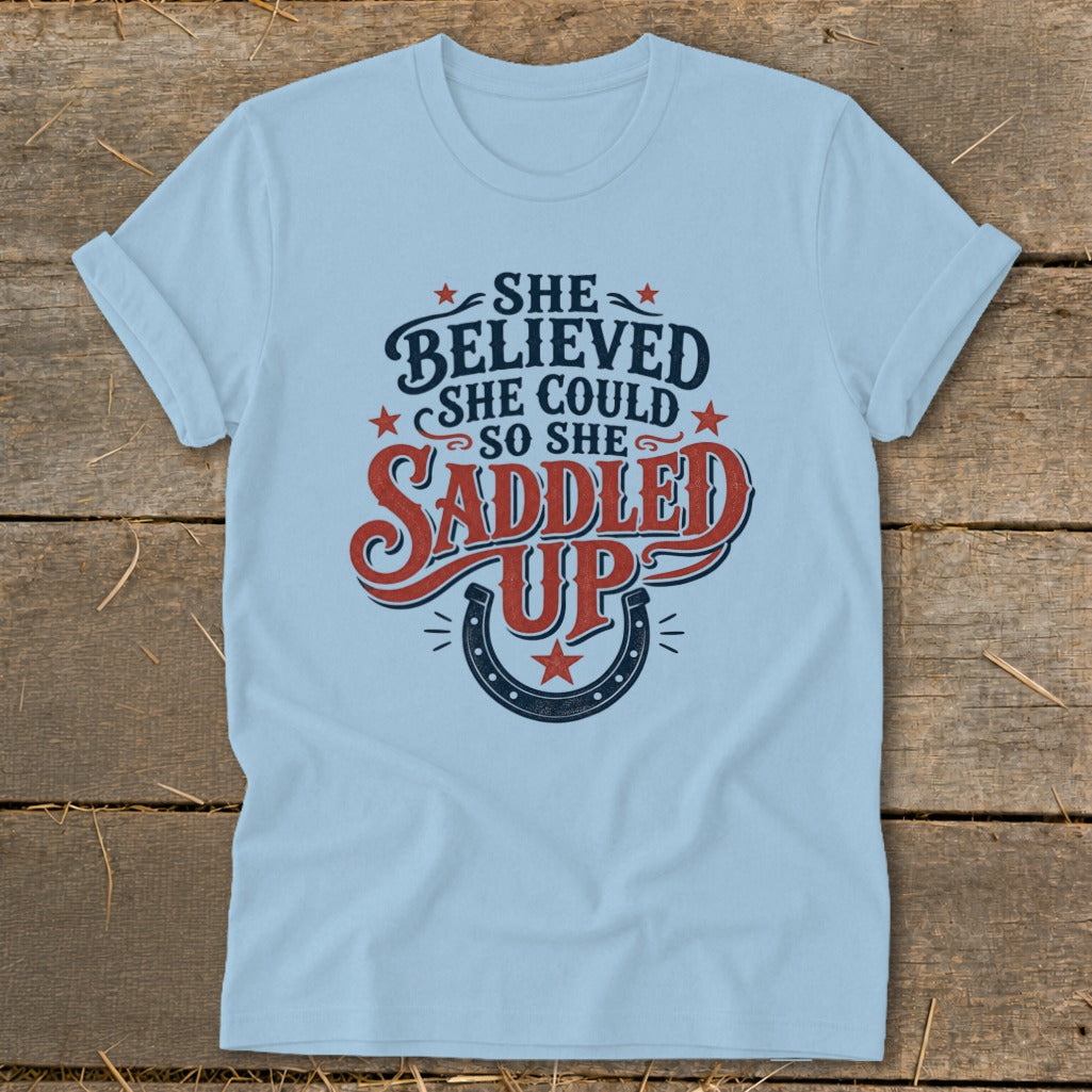 Light blue t-shirt with "She Believed She Could So She Saddled Up" text and horseshoe.