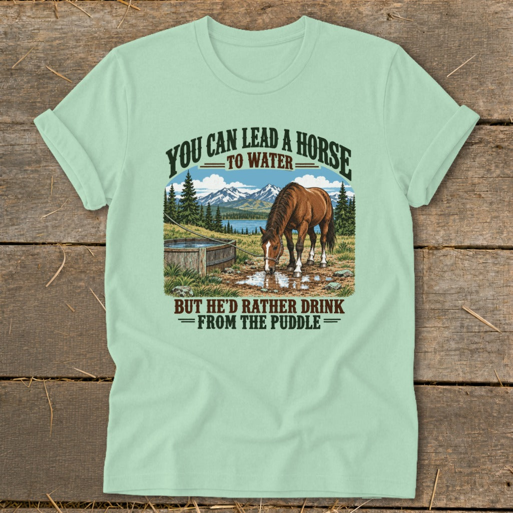 A light green t-shirt with a vintage illustration of a horse drinking from a puddle, featuring the text you can lead a horse to water, but he'd rather drink from the puddle.