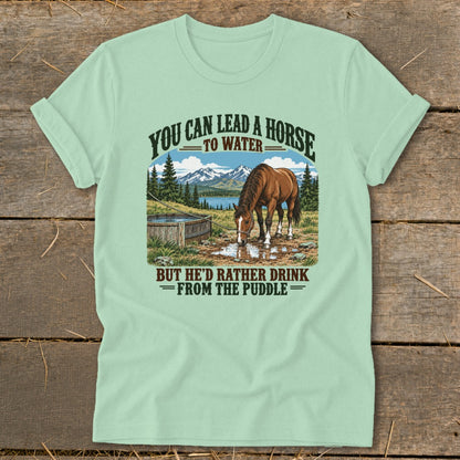 A light green t-shirt with a vintage illustration of a horse drinking from a puddle, featuring the text you can lead a horse to water, but he'd rather drink from the puddle.