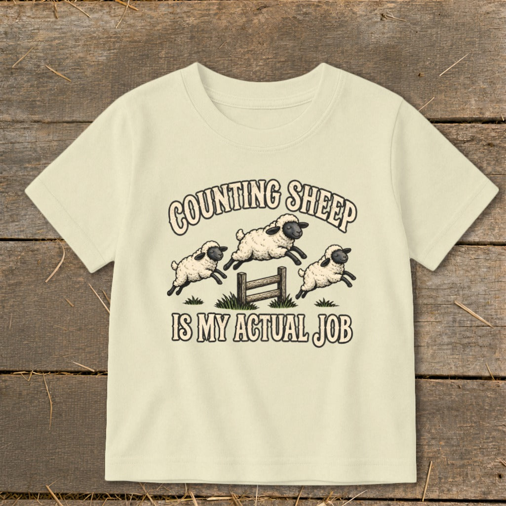Cream t-shirt with cartoon sheep jumping over a fence and text "Counting Sheep Is My Actual Job."